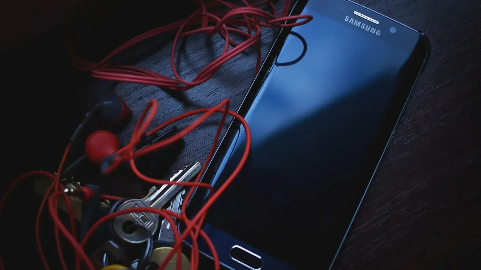 Smartphone Samsung con auriculares rojos y llaves sobre una mesa de madera, en iluminación natural.