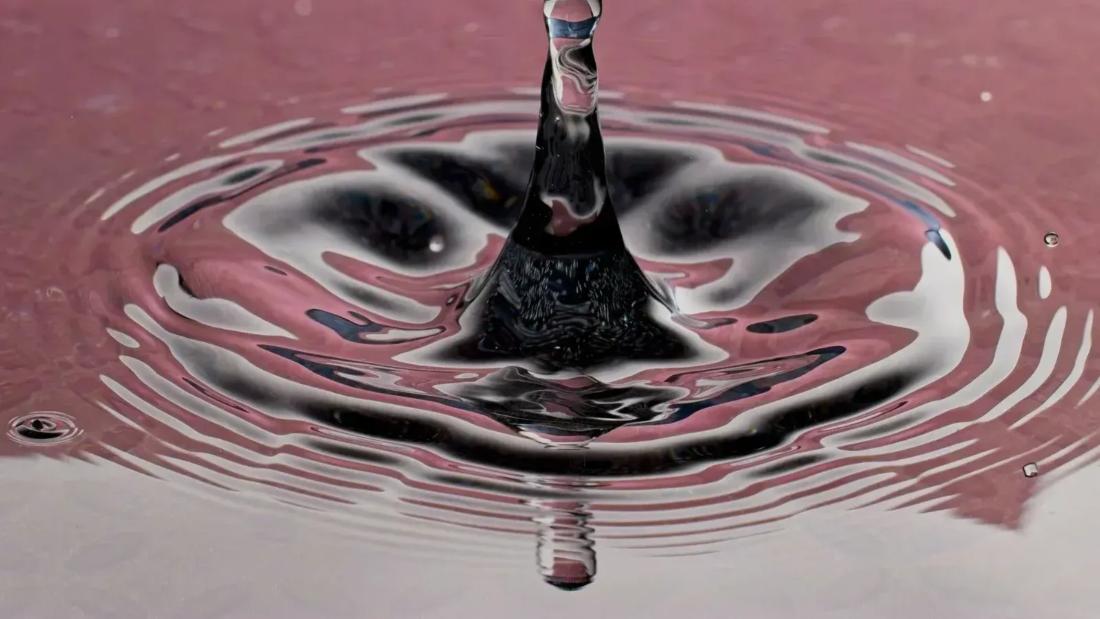 Cautivante salpicadura de gotas de agua creando ondas en una superficie rosa, mostrando la belleza de la dinámica de fluidos.