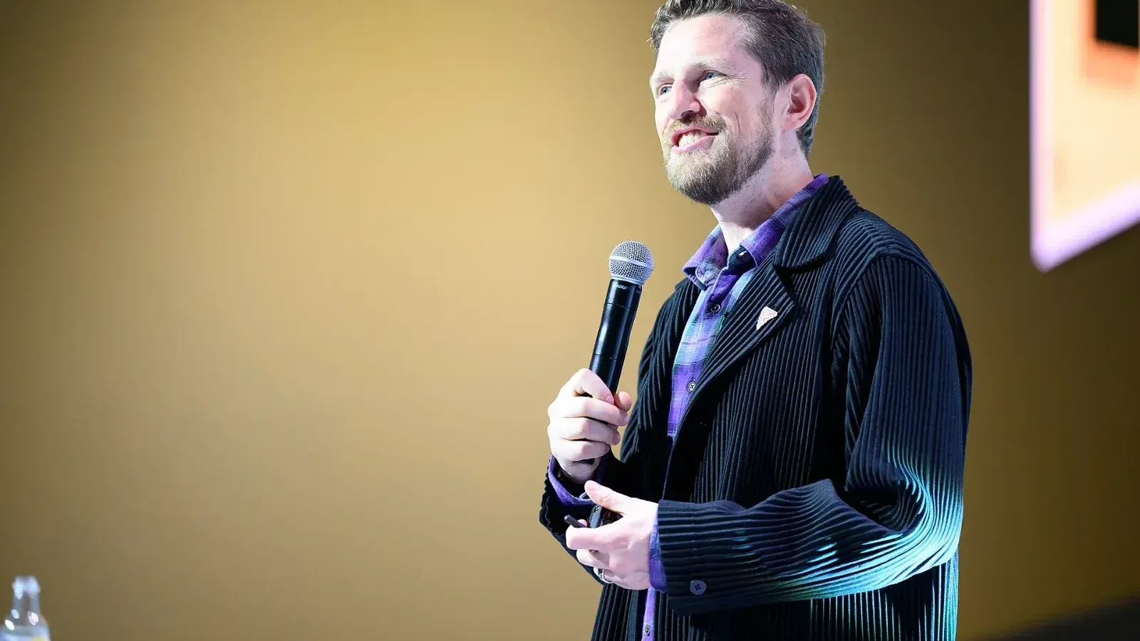 Matt Mullenweg habla sonriente con un micrófono en la mano sobre un escenario, con saco negro y camisa morada a cuadros.