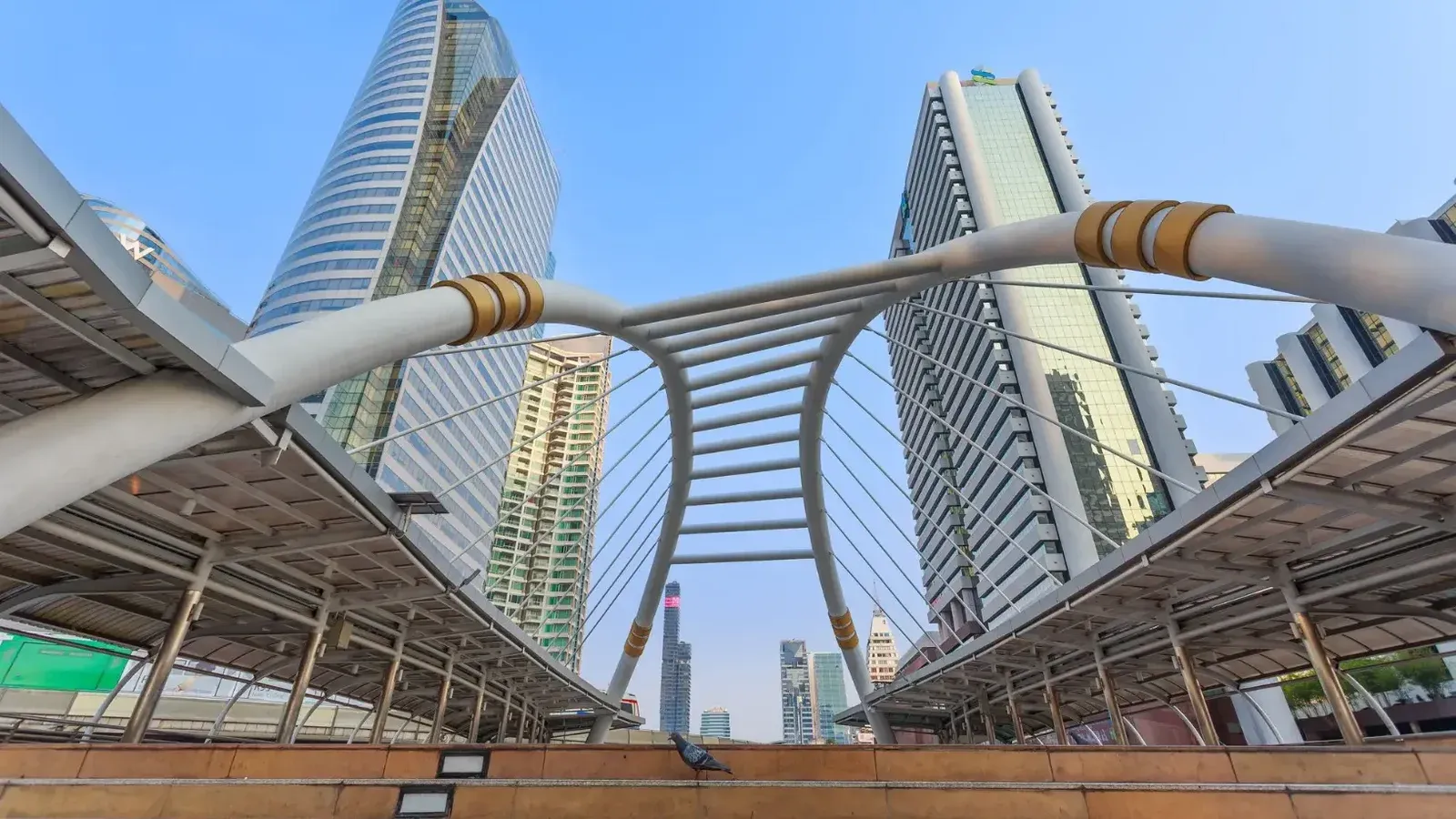 Vista dinámica del horizonte de Bangkok con imponentes rascacielos y el icónico puente Skywalk bajo un cielo azul claro.