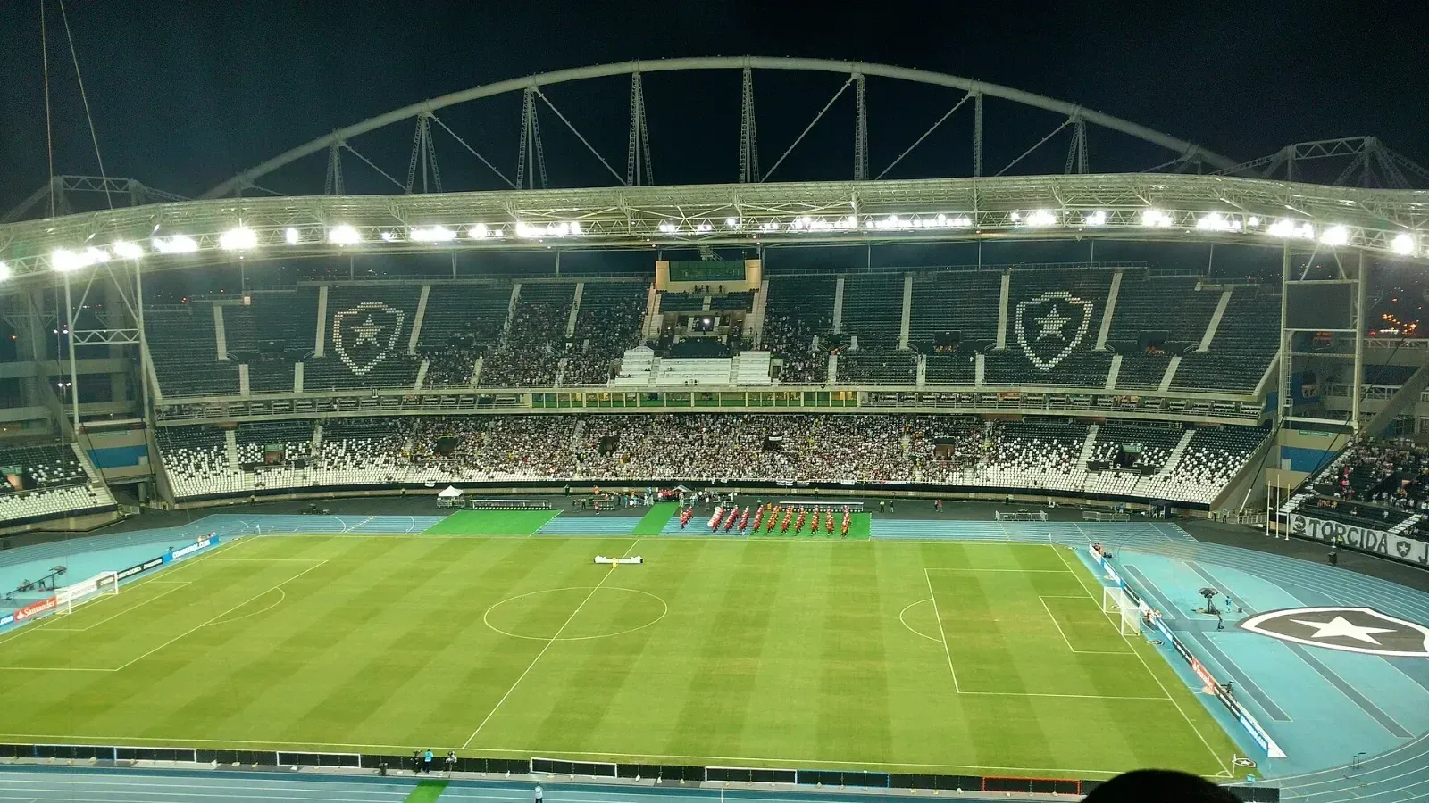 Vista nocturna del Estádio Nilton Santos, en Río de Janeiro, con escudos del Botafogo en las gradas y un grupo de personas vestidas de rojo junto al césped antes de un partido