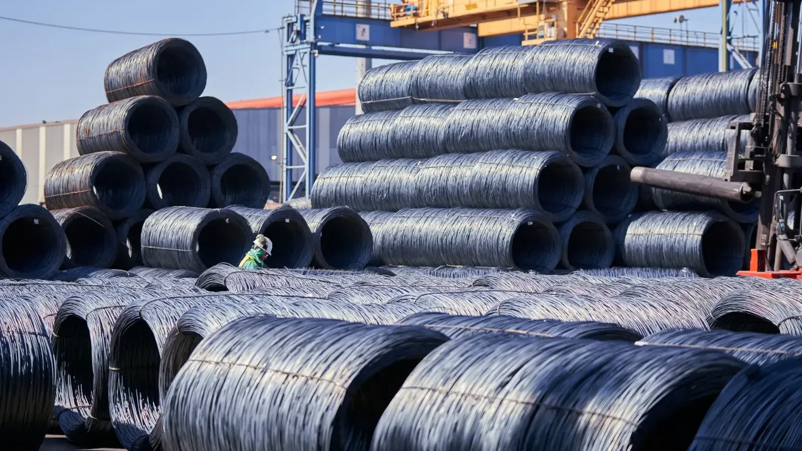 Grandes pilas de bobinas de alambre de acero almacenadas al aire libre en un sitio industrial bajo la luz del día.