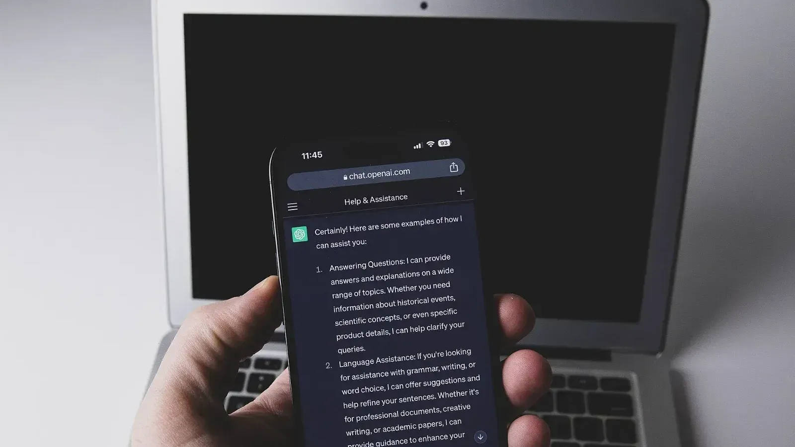 Mano que sostiene un teléfono inteligente con la interfaz de ChatGPT abierta en chat.openai.com frente a una laptop con la pantalla apagada