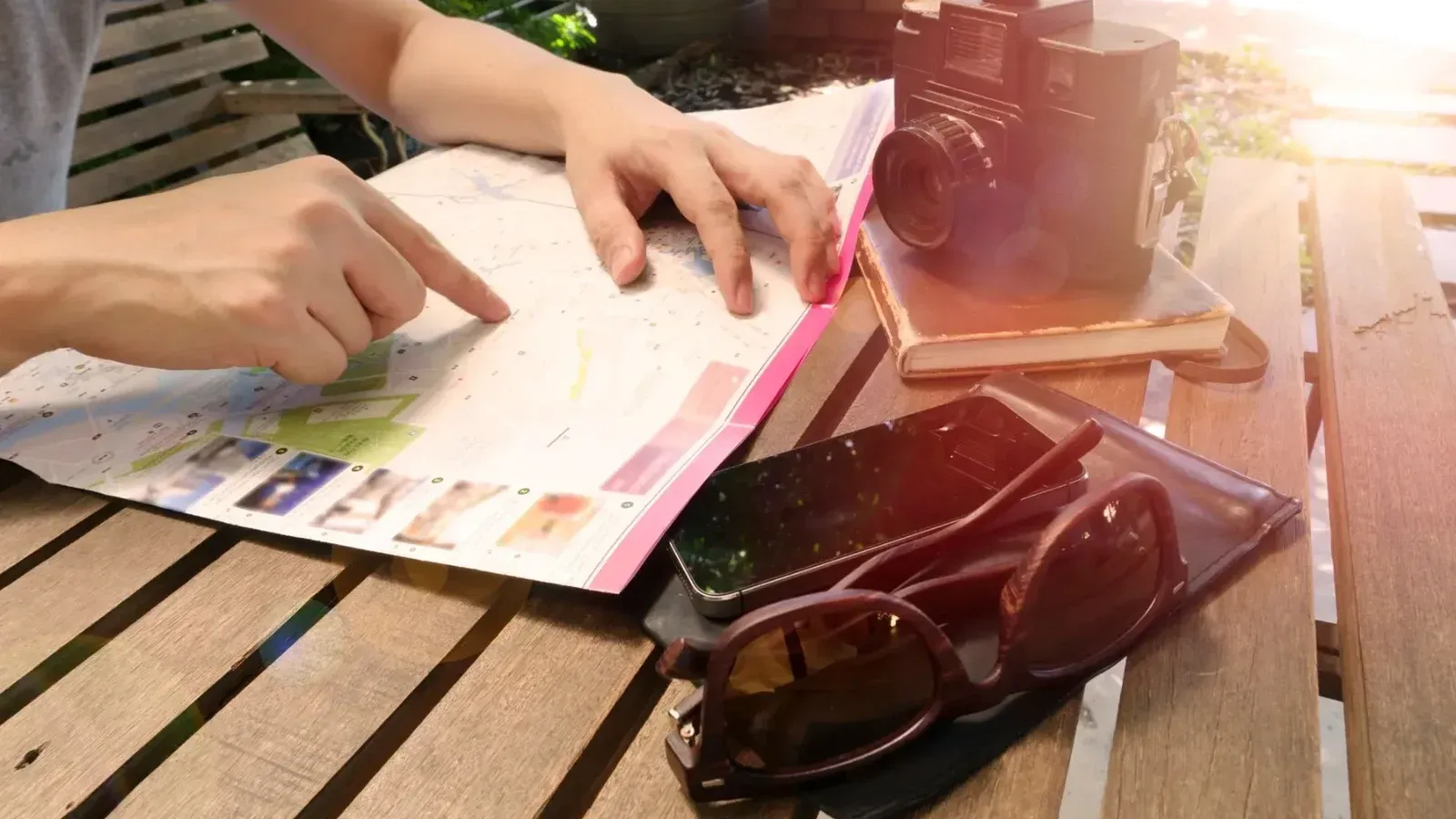 Viajero apuntando al mapa sobre mesa de madera con cámara y accesorios bajo la luz del sol.