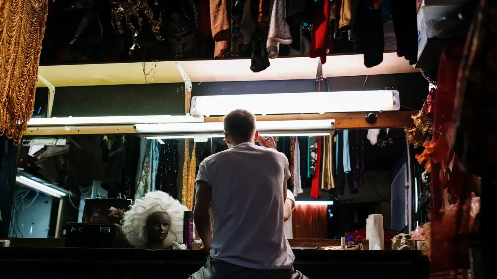 Una persona anónima preparando entre bastidores con disfraces, pelucas y espejos en un vestidor.