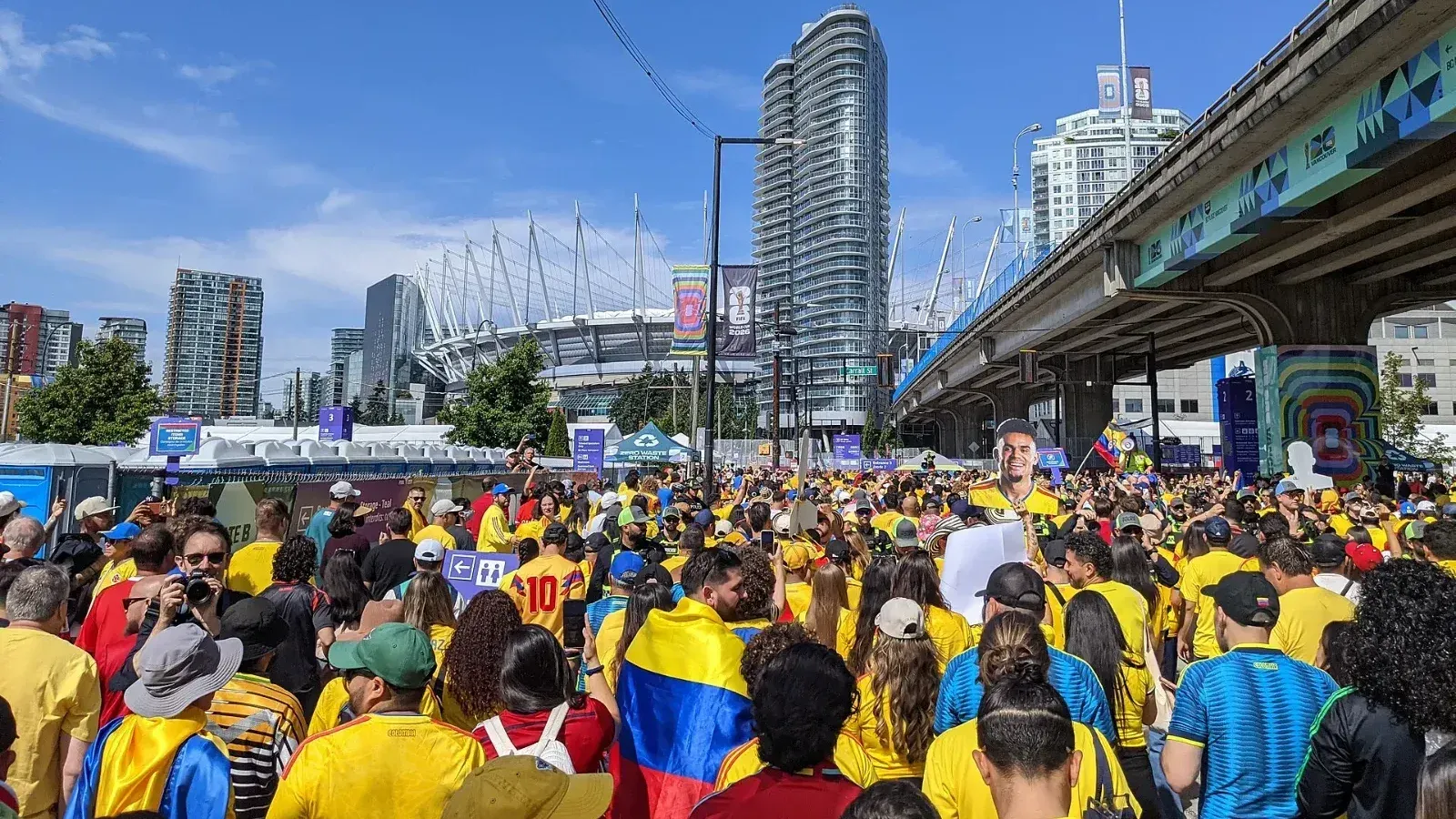 Multitud de aficionados de Colombia con camisetas amarillas y banderas tricolores camina por una calle hacia el estadio BC Place de Vancouver, con cielo despejado.