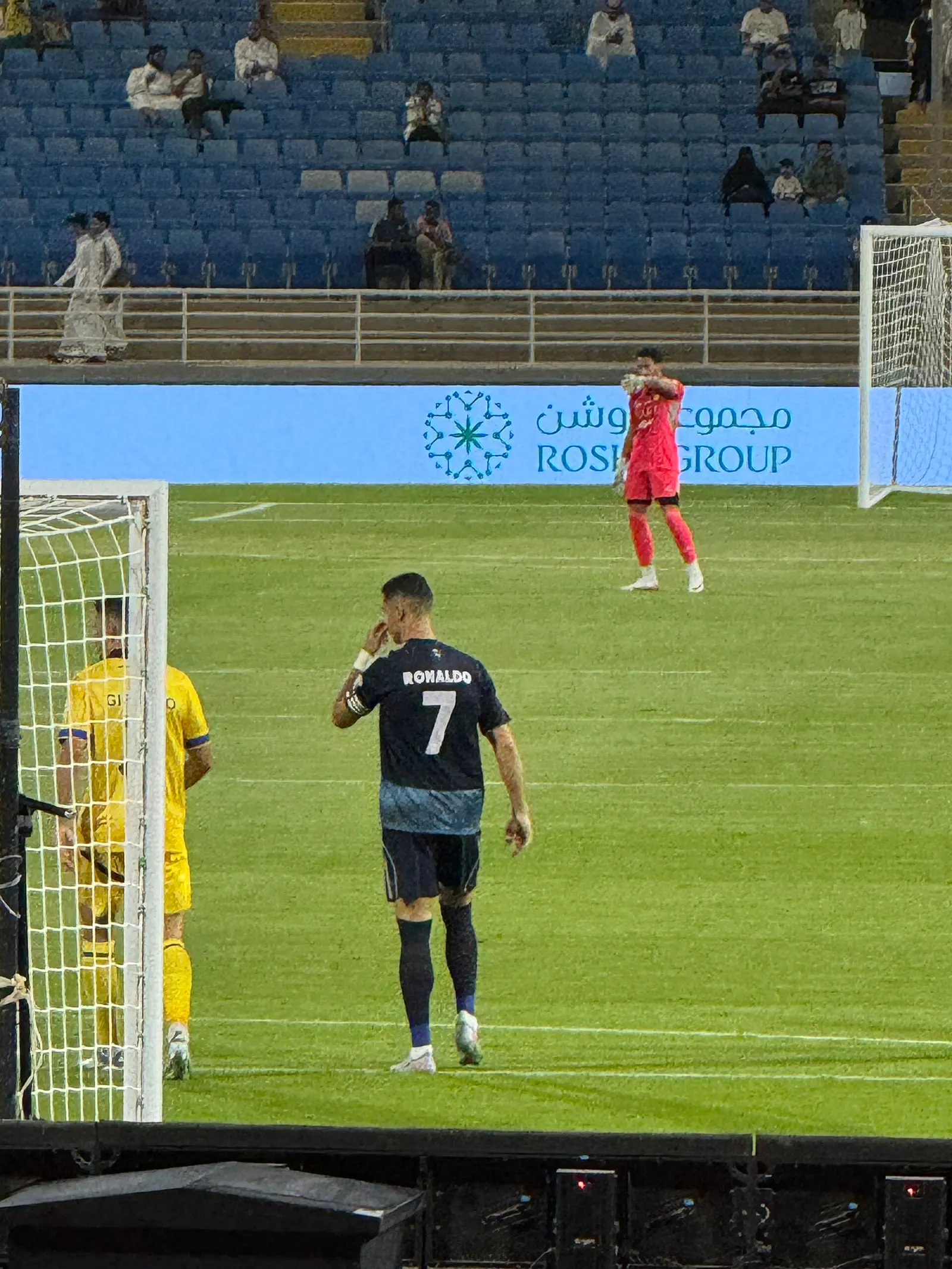 Cristiano Ronaldo, de espaldas con la camiseta oscura número 7, camina por el césped junto a una portería; al fondo, un portero de rojo y gradas azules casi vacías.