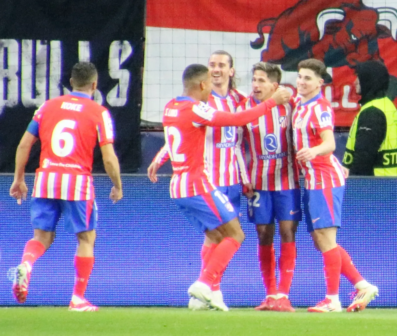 Cuatro futbolistas del Atlético de Madrid con camiseta rojiblanca se abrazan sonrientes sobre el césped mientras un quinto se acerca, junto a una valla publicitaria.