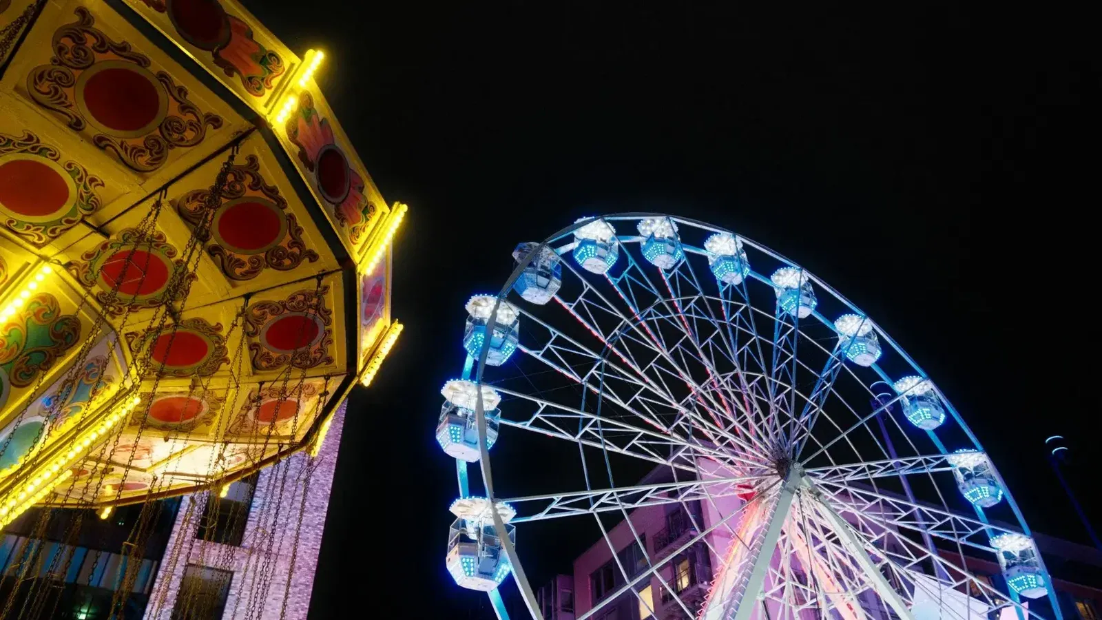 A vibrant Ferris wheel and spinning ride lit up at a night carnival, creating a lively festive atmosphere.
