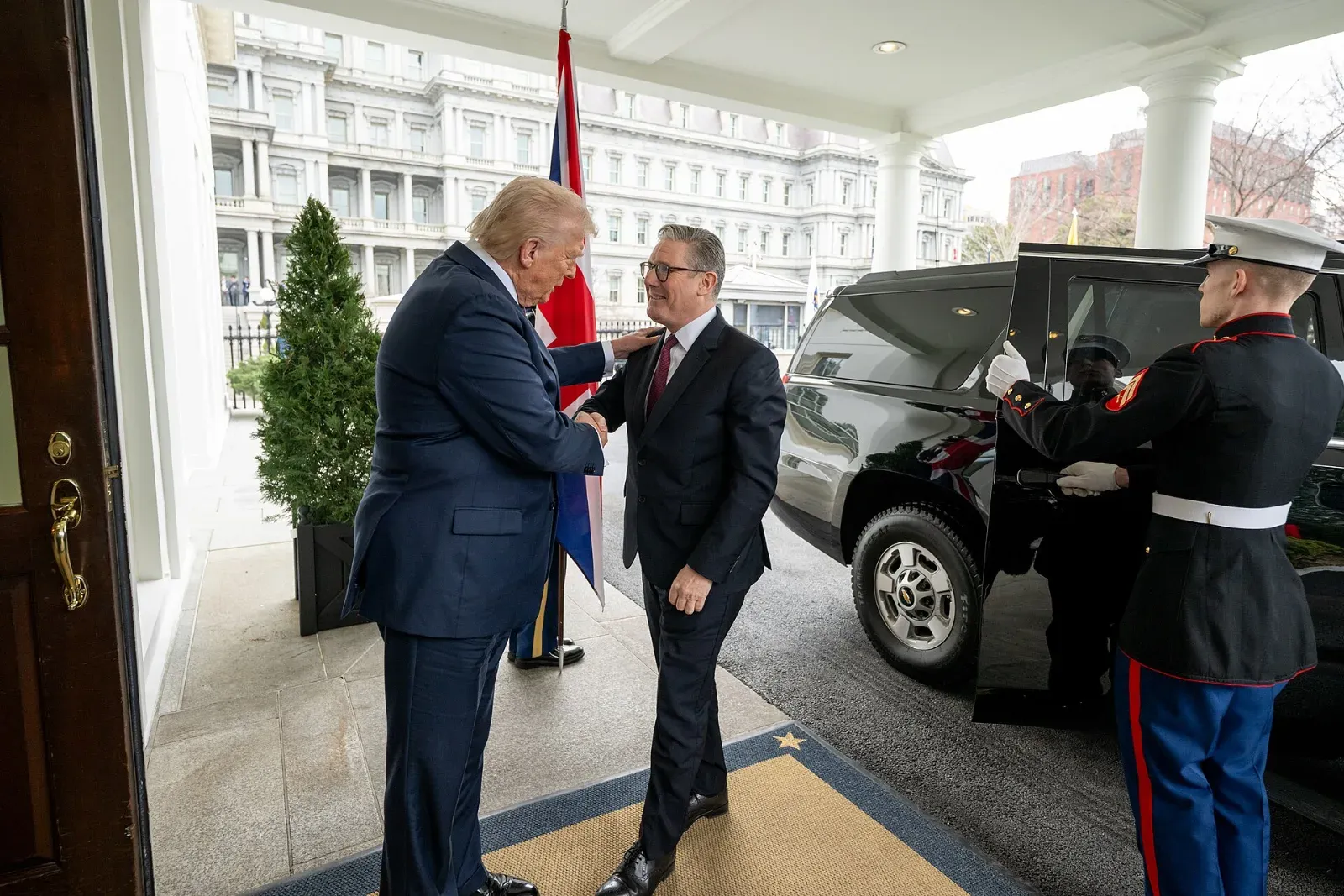 Donald Trump estrecha la mano de Keir Starmer en una entrada de la Casa Blanca, junto a una bandera británica y un marine que abre la puerta de un vehículo.