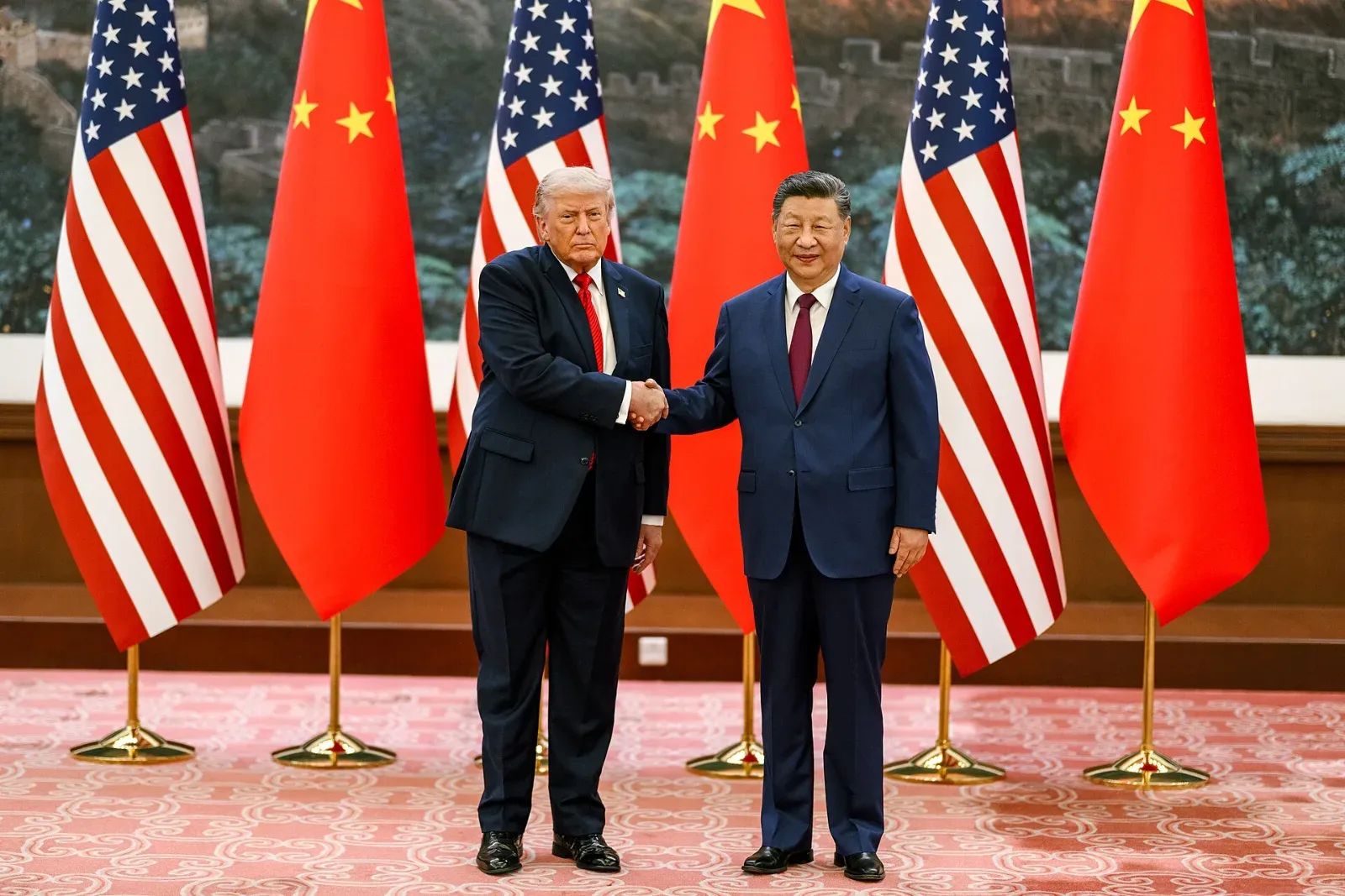 Donald Trump y Xi Jinping se dan la mano frente a banderas alternadas de Estados Unidos y China, de pie sobre una alfombra roja.