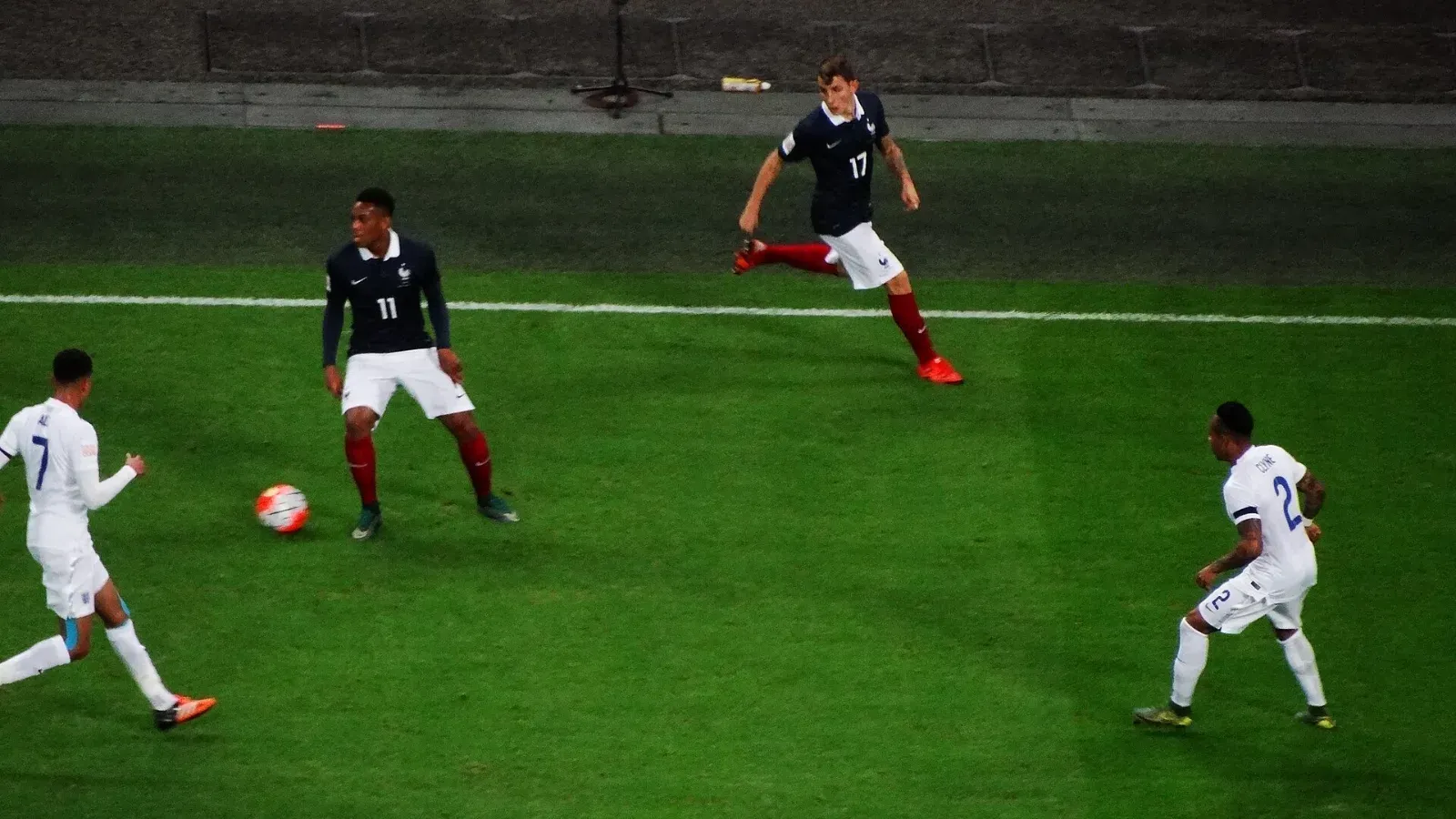 Dos futbolistas de Francia con camiseta azul marino y dos de Inglaterra con camiseta blanca se mueven alrededor del balón sobre el césped.