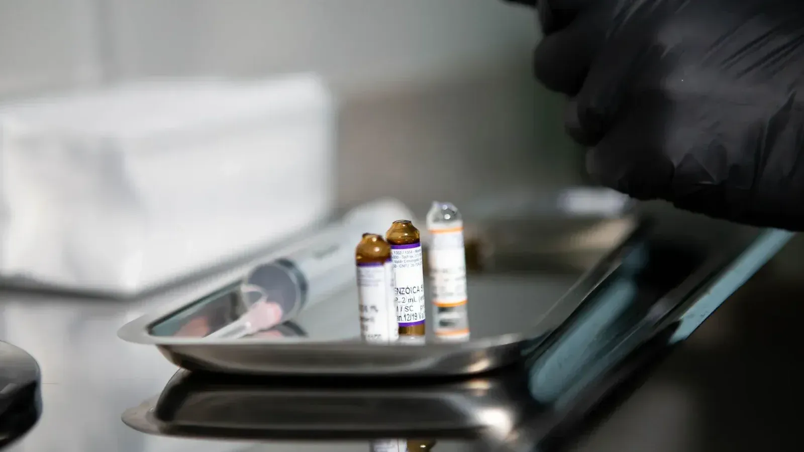 Primer plano de viales médicos y jeringa en bandeja de acero inoxidable en entorno clínico.