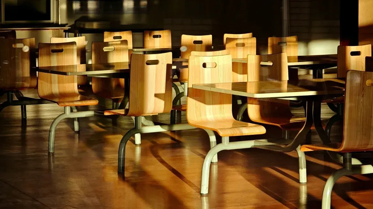 Sillas y mesas de madera vacías en un entorno de cafetería iluminado por el sol, proyectando sombras dramáticas.