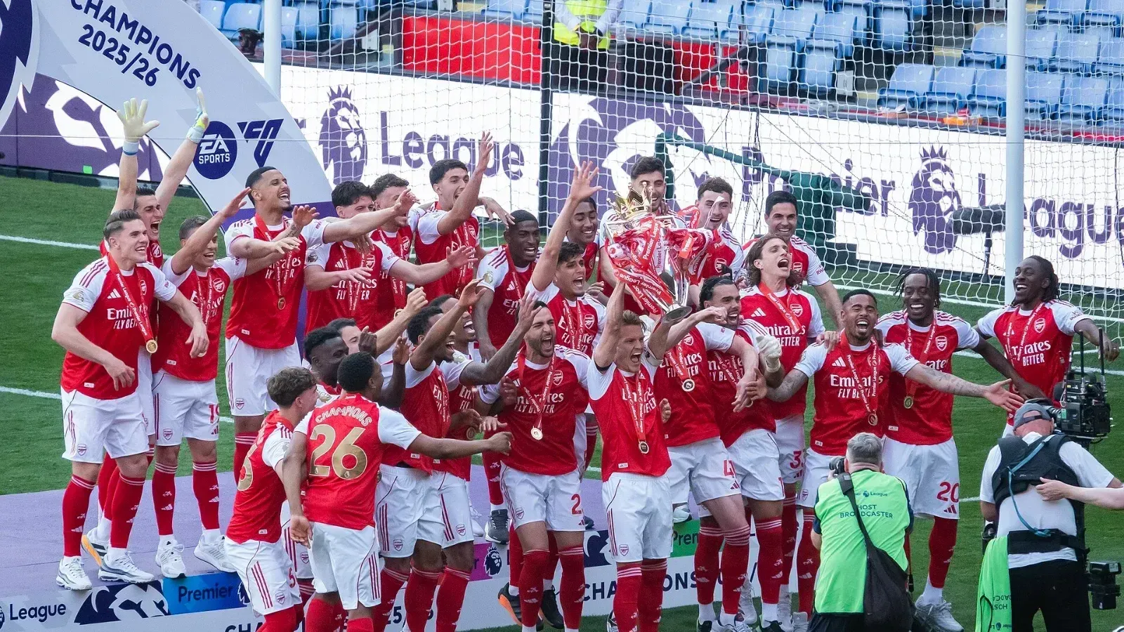 Jugadores del Arsenal, con medallas al cuello, celebran sobre el césped alrededor del trofeo de la Premier League ante carteles de campeones 2025/26.