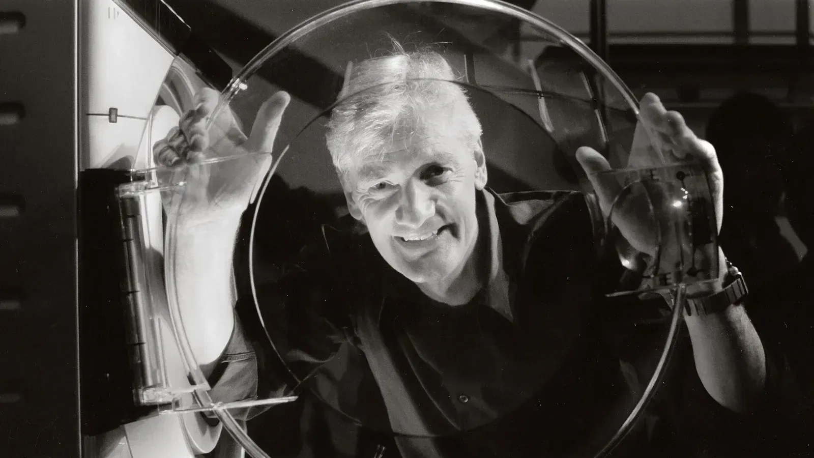 James Dyson sonríe mientras sostiene con ambas manos la puerta transparente de una lavadora, en una fotografía en blanco y negro.