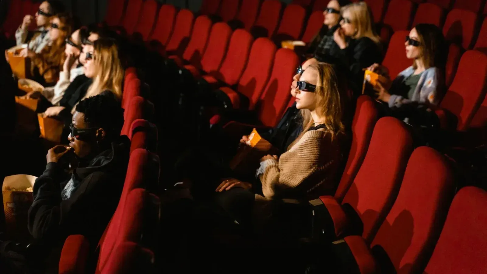 Gente con gafas 3D y comiendo palomitas en un cine con sillas rojas.