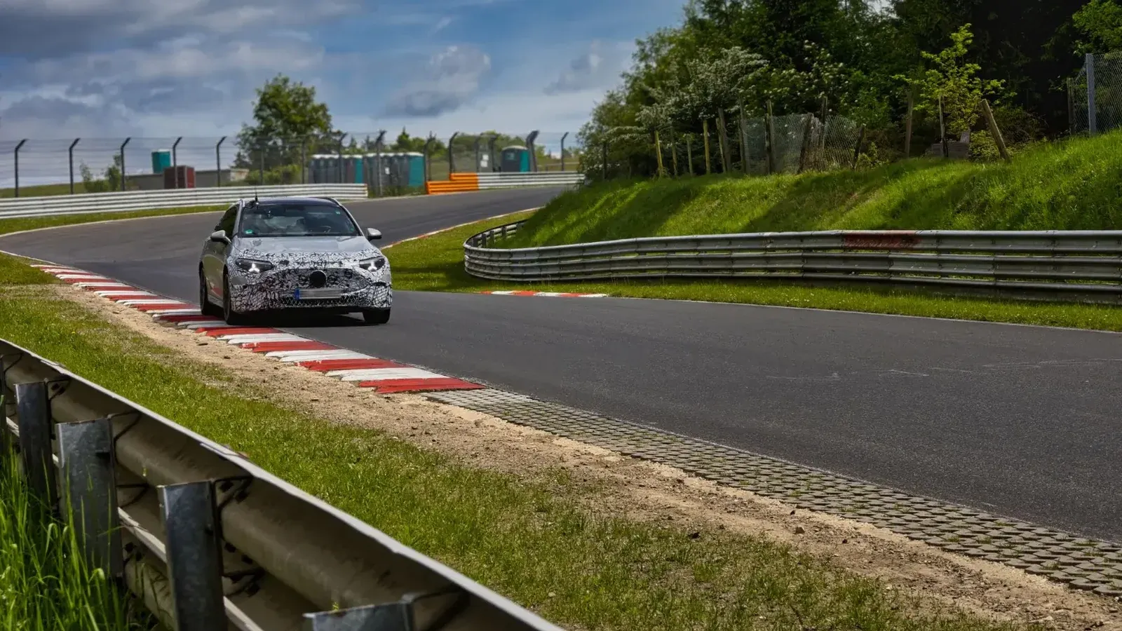 A camouflaged sports car speeds around the Nürburgring race track on a sunny day.