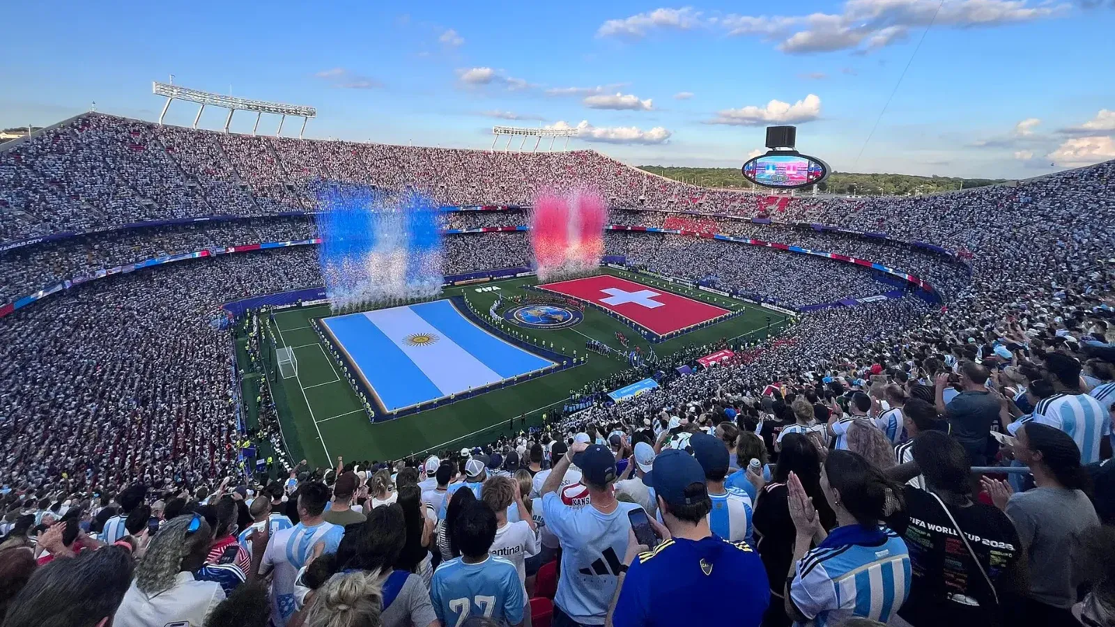 Vista panorámica de un estadio lleno, con grandes banderas de Argentina y Suiza desplegadas sobre el césped y humo azul, blanco y rojo antes del partido.