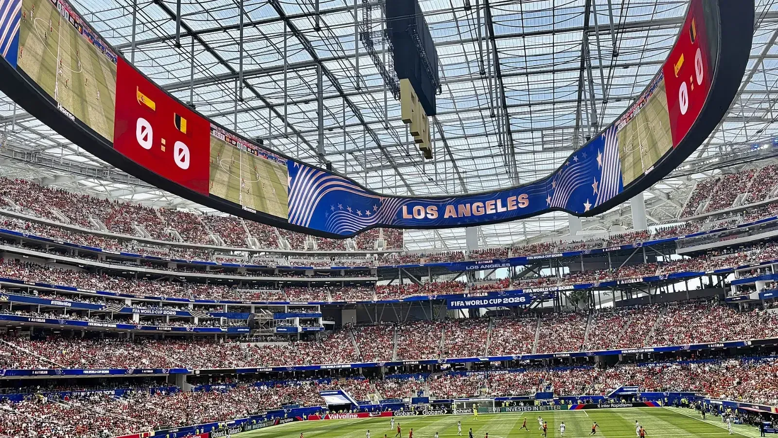 Estadio lleno visto desde la grada baja durante un partido de fútbol, con el videomarcador en 0-0 junto a las banderas de España y Bélgica y el rótulo Los Angeles.