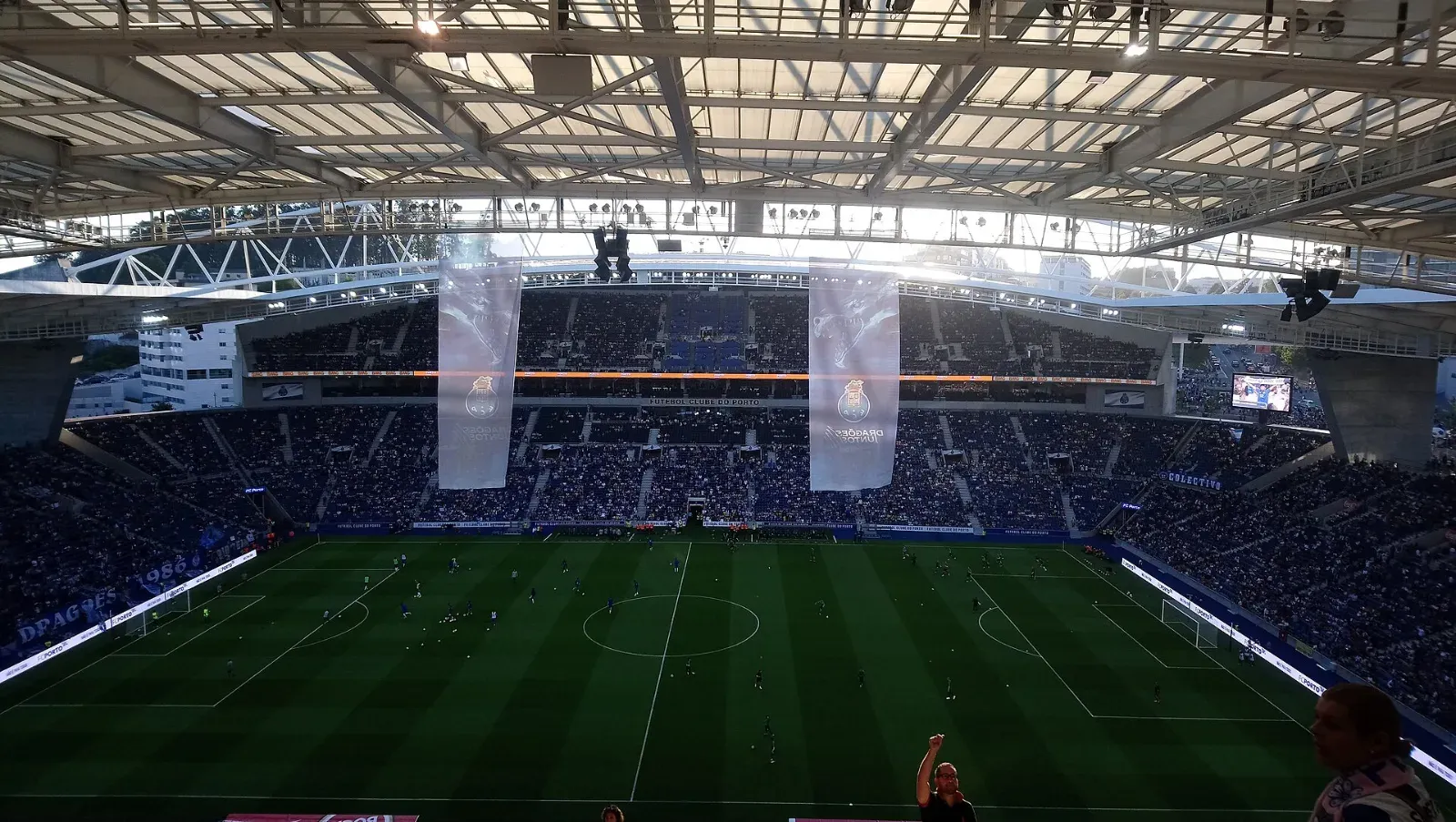 Estadio de fútbol con las gradas llenas bajo un techo iluminado por el sol; dos grandes estandartes con el escudo del FC Porto cuelgan del techo frente a la tribuna.