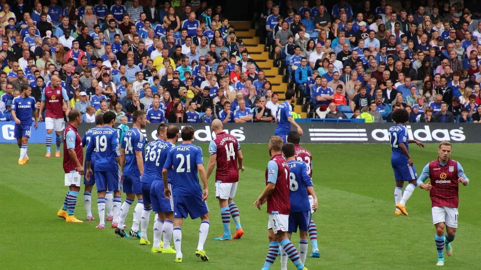 Jugadores del Chelsea, de azul, y del Aston Villa, de granate y celeste, esperan una jugada a balón parado frente a una grada llena de aficionados.