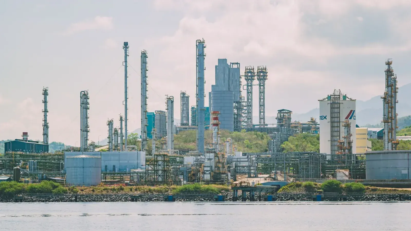 Un gran complejo de refinería industrial con chimeneas altas junto al mar bajo un cielo despejado.