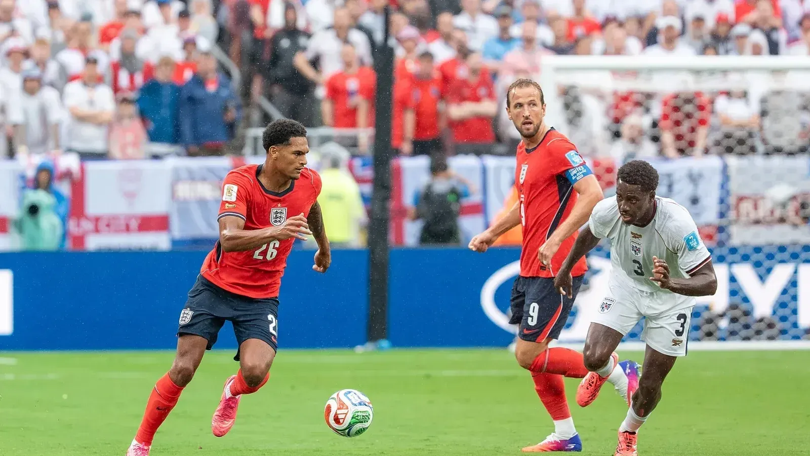 Jarell Quansah, con la camiseta roja de Inglaterra y el número 26, conduce el balón; detrás corren un compañero con el 9 y un rival de camiseta blanca.