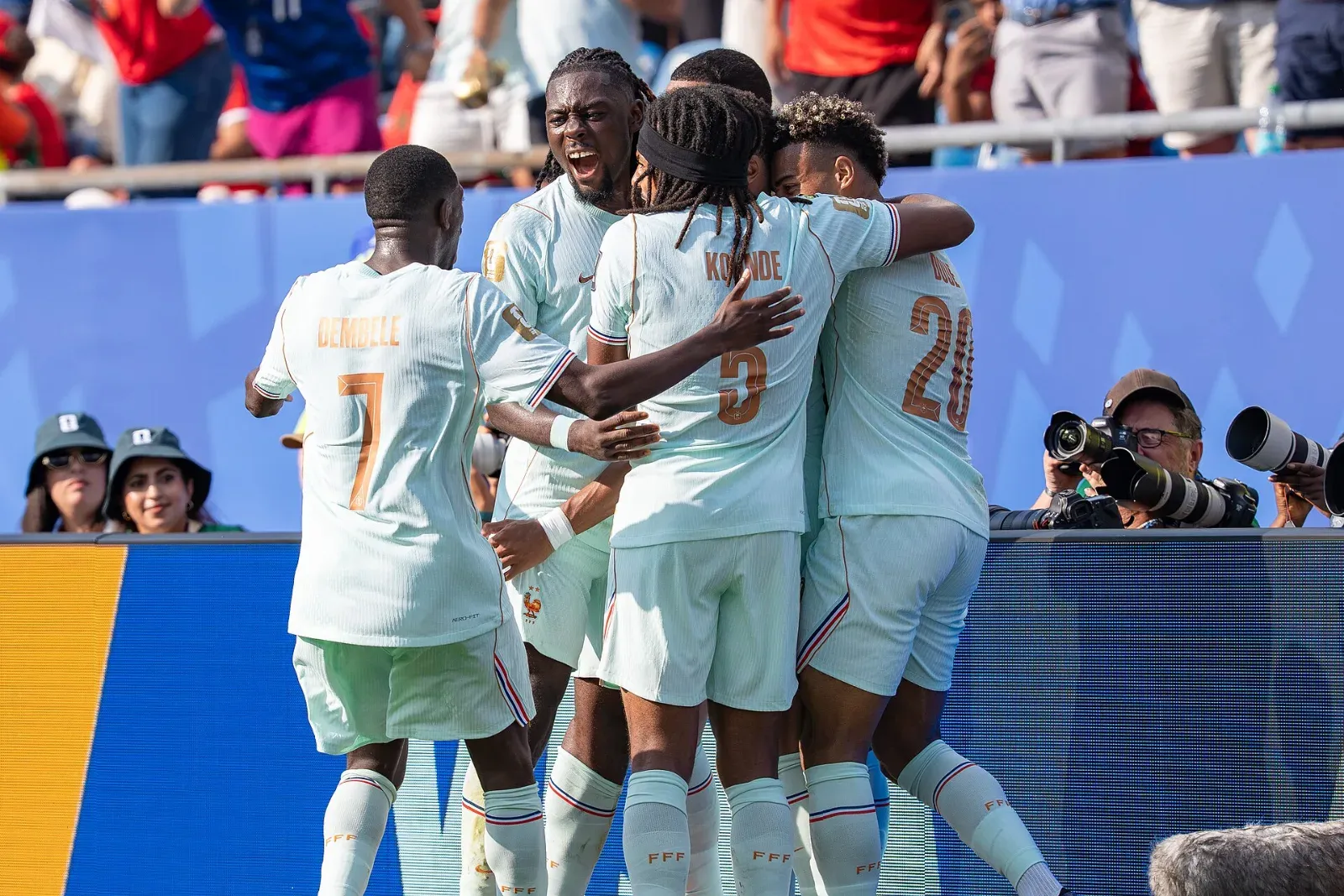 Futbolistas de Francia con camiseta blanca se abrazan para celebrar junto a la banda, con fotógrafos y aficionados detrás.