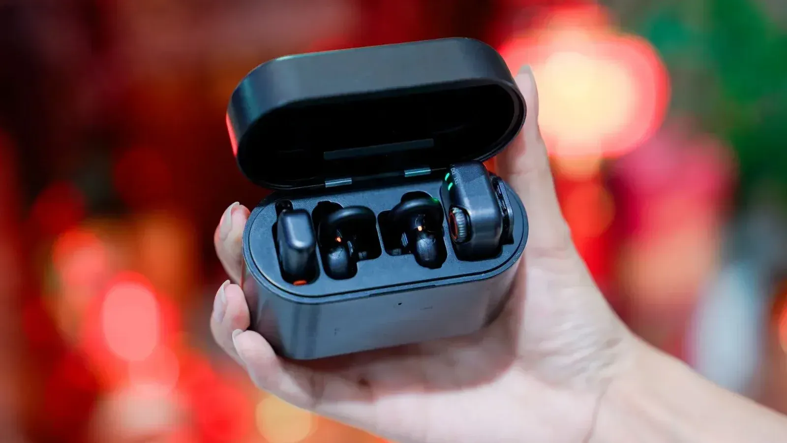 A hand holds a pair of black wireless earbuds in an open charging case.
