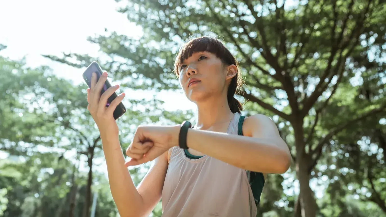 Desde abajo étnica mujer asiática con ropa deportiva de pie en el parque y conectando smartphone y reloj inteligente durante el descanso en día soleado