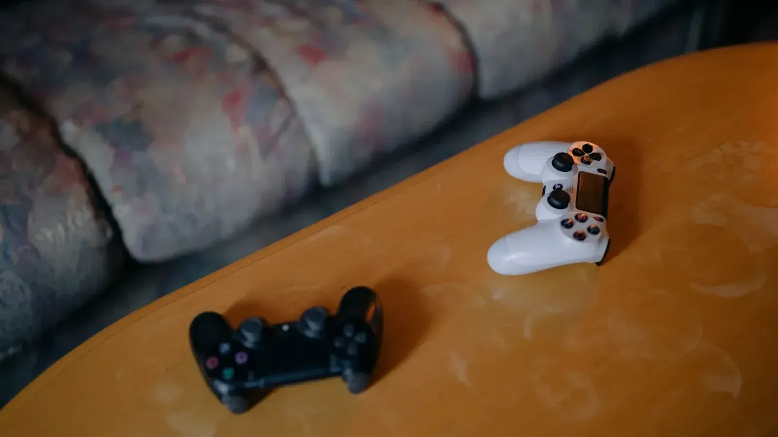 Controladores de juego en blanco y negro colocados sobre una mesa de madera con un sofá borroso en el fondo.