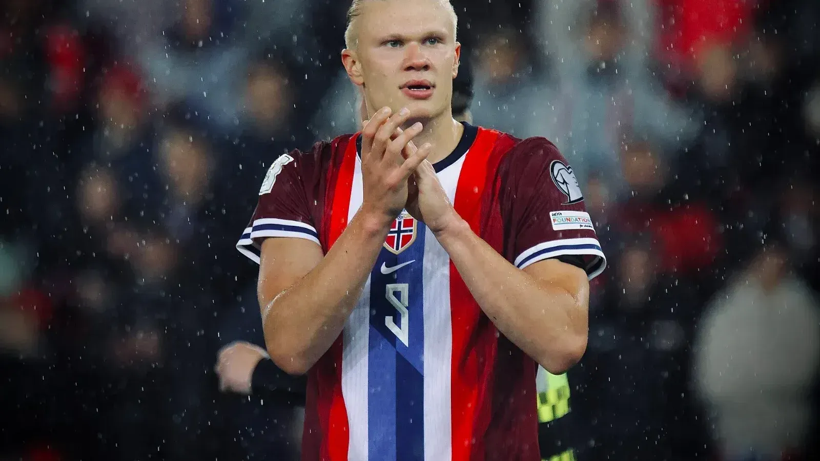 Erling Haaland, con la camiseta roja de Noruega y el número 9, aplaude con las manos juntas bajo la lluvia frente a una grada de aficionados.