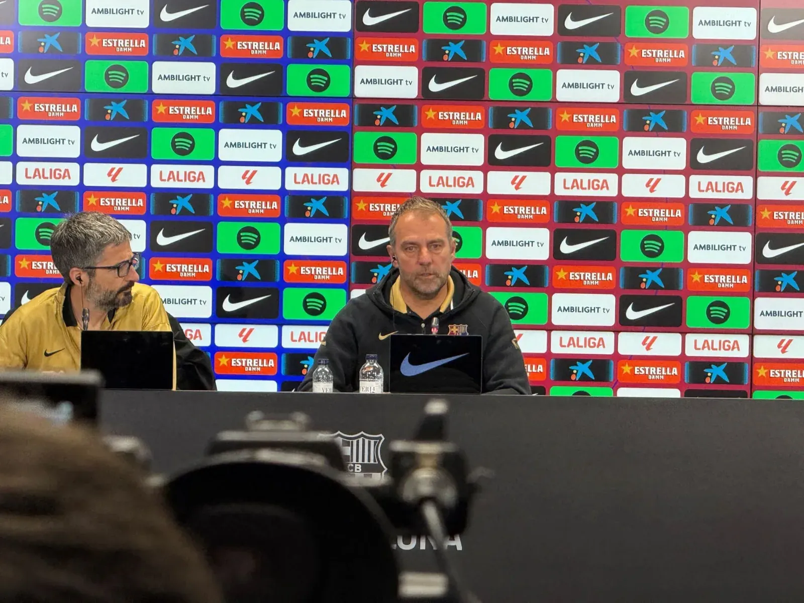 Hansi Flick, con sudadera oscura del FC Barcelona, habla en la mesa de una rueda de prensa frente a un panel de patrocinadores; a su lado, un hombre con gafas.
