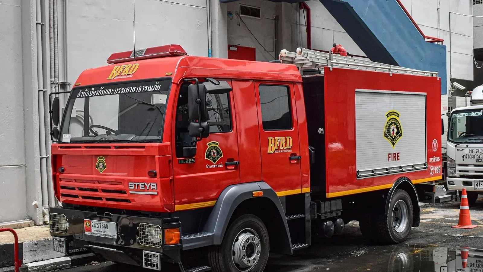 Camión de bomberos rojo del Departamento de Bomberos y Rescate de Bangkok (BFRD), de la marca Steyr, estacionado sobre pavimento mojado junto a un edificio.