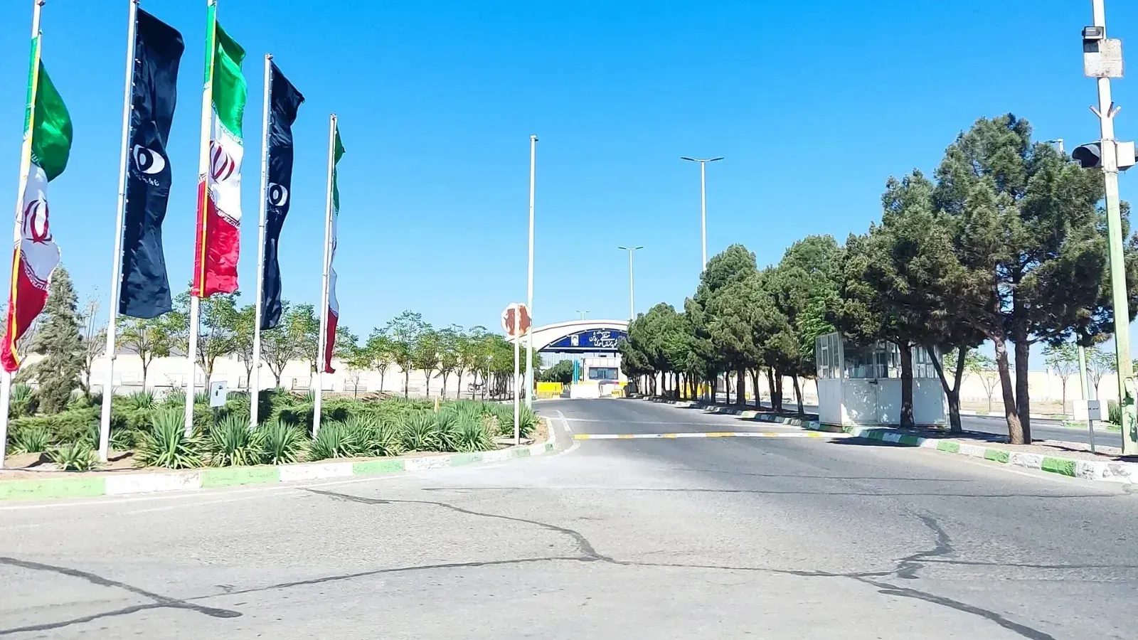 Carretera de acceso a la instalación nuclear de Natanz, con banderas de Irán y banderas azul oscuro con un emblema junto a la vía, árboles a un costado y un pórtico de entrada al fondo.