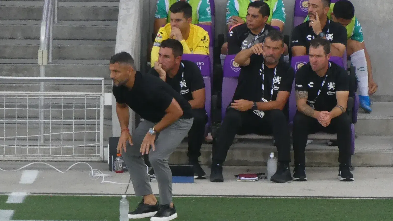 Javier Gandolfi, inclinado con una mano sobre la rodilla, sigue un partido al borde de la cancha junto al cuerpo técnico del Club León sentado en la banca.