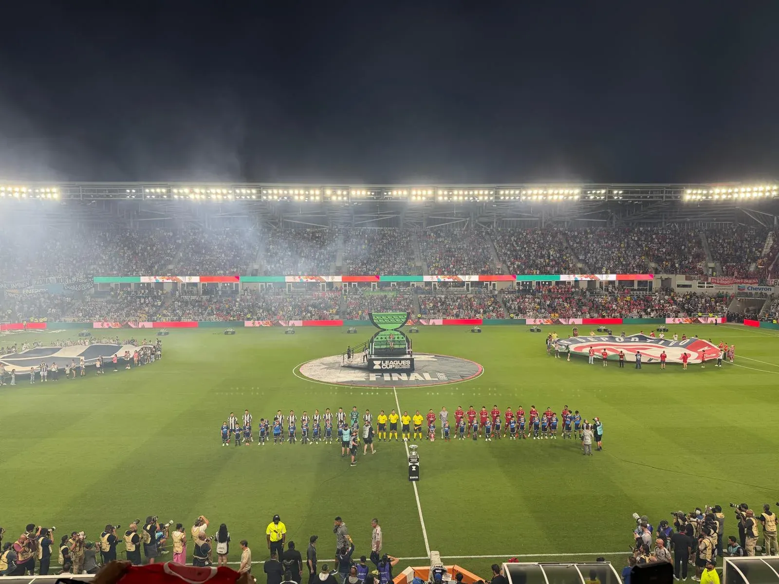 Jugadores de dos equipos, con uniformes rojos y de rayas, formados en el centro del campo junto al logotipo de la final de la Leagues Cup, en un estadio lleno de noche.