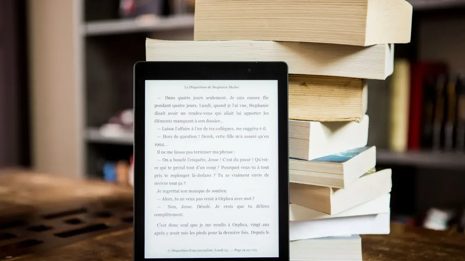 E-reader with text displayed next to a stack of physical books on a wooden table.