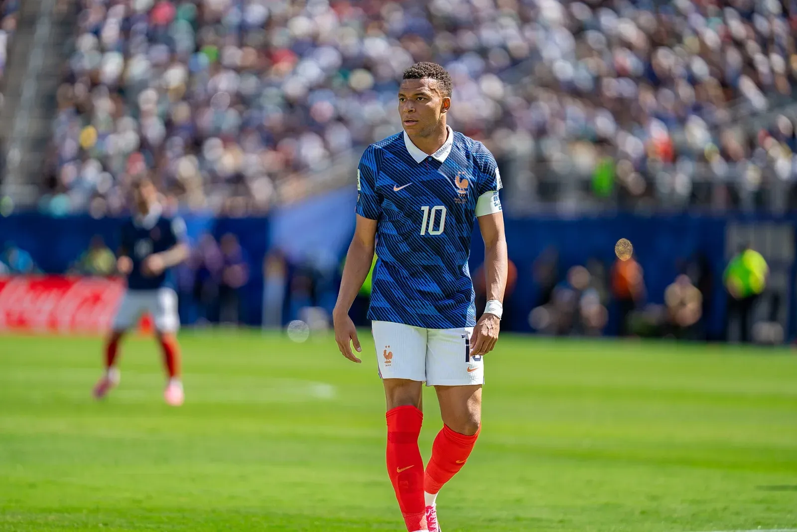 Kylian Mbappé, con la camiseta azul número 10 de Francia, medias rojas y brazalete de capitán, camina sobre el césped con la grada llena al fondo.