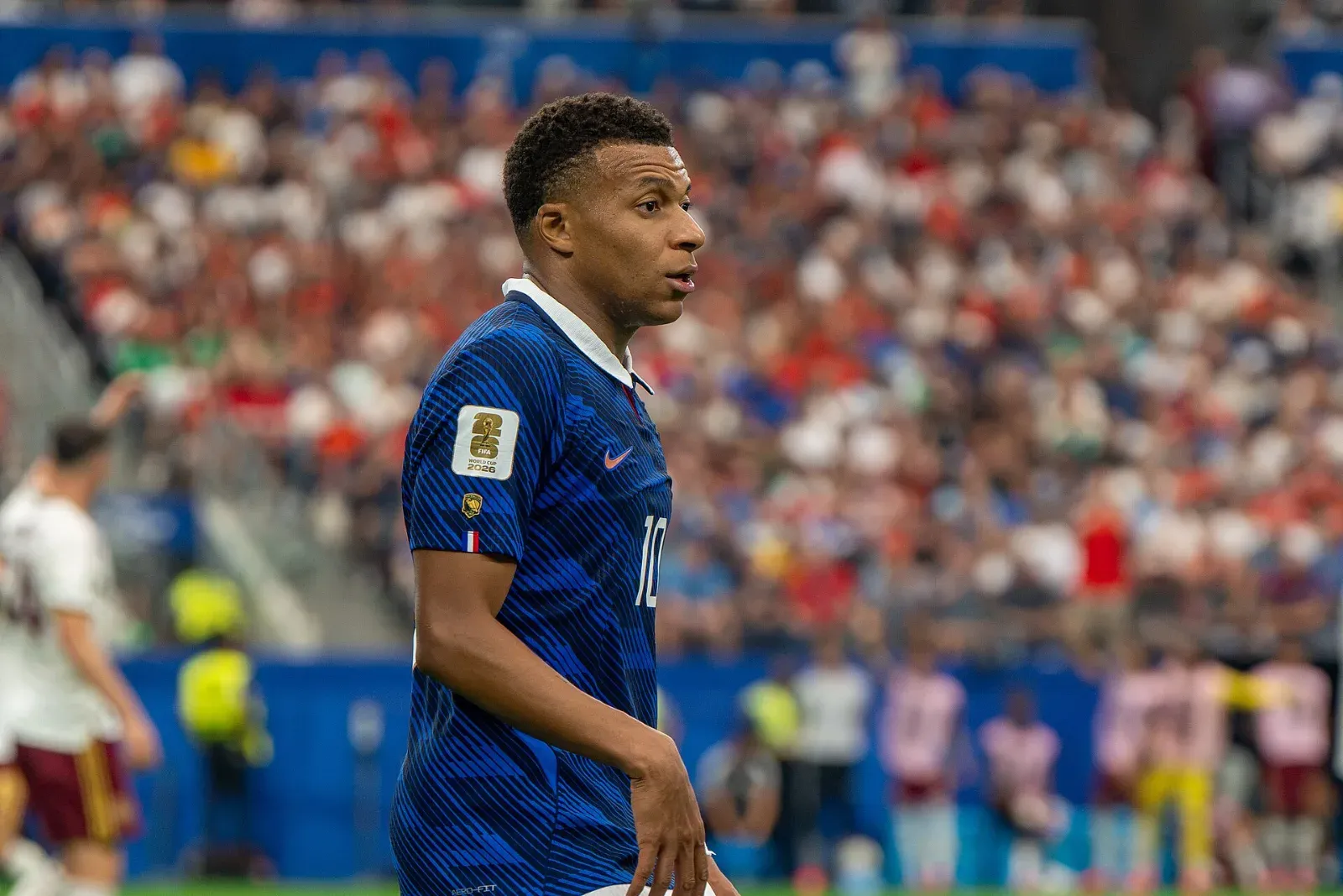 Kylian Mbappé, con la camiseta azul número 10 de Francia, mira hacia un lado sobre el campo con la grada llena al fondo.