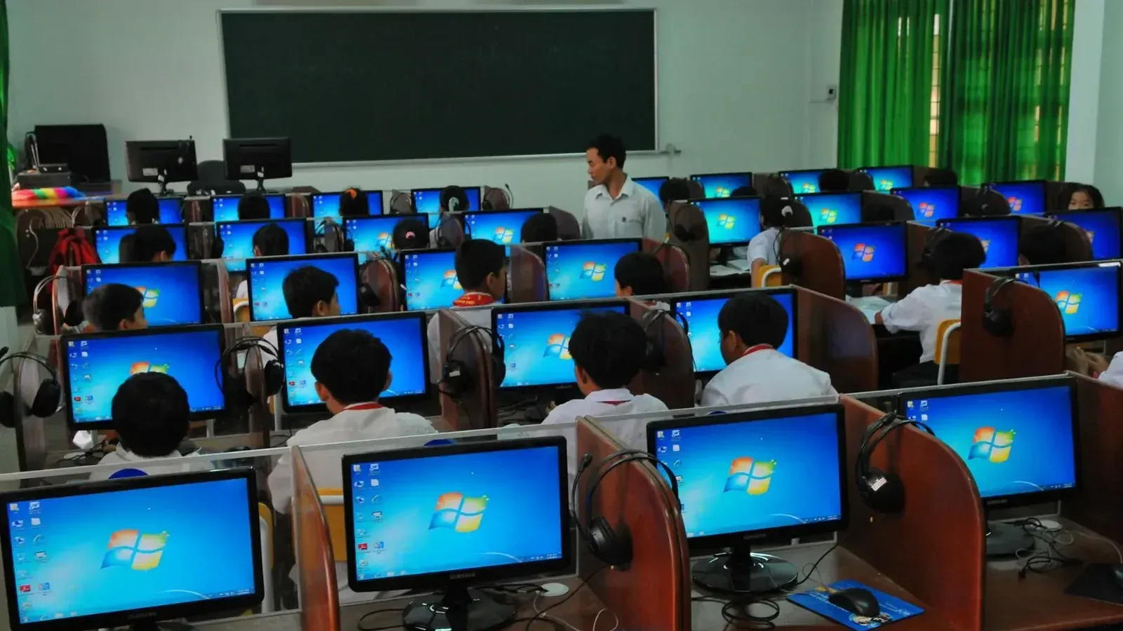 Students learning in a computer lab with multiple monitors and a teacher instructing.