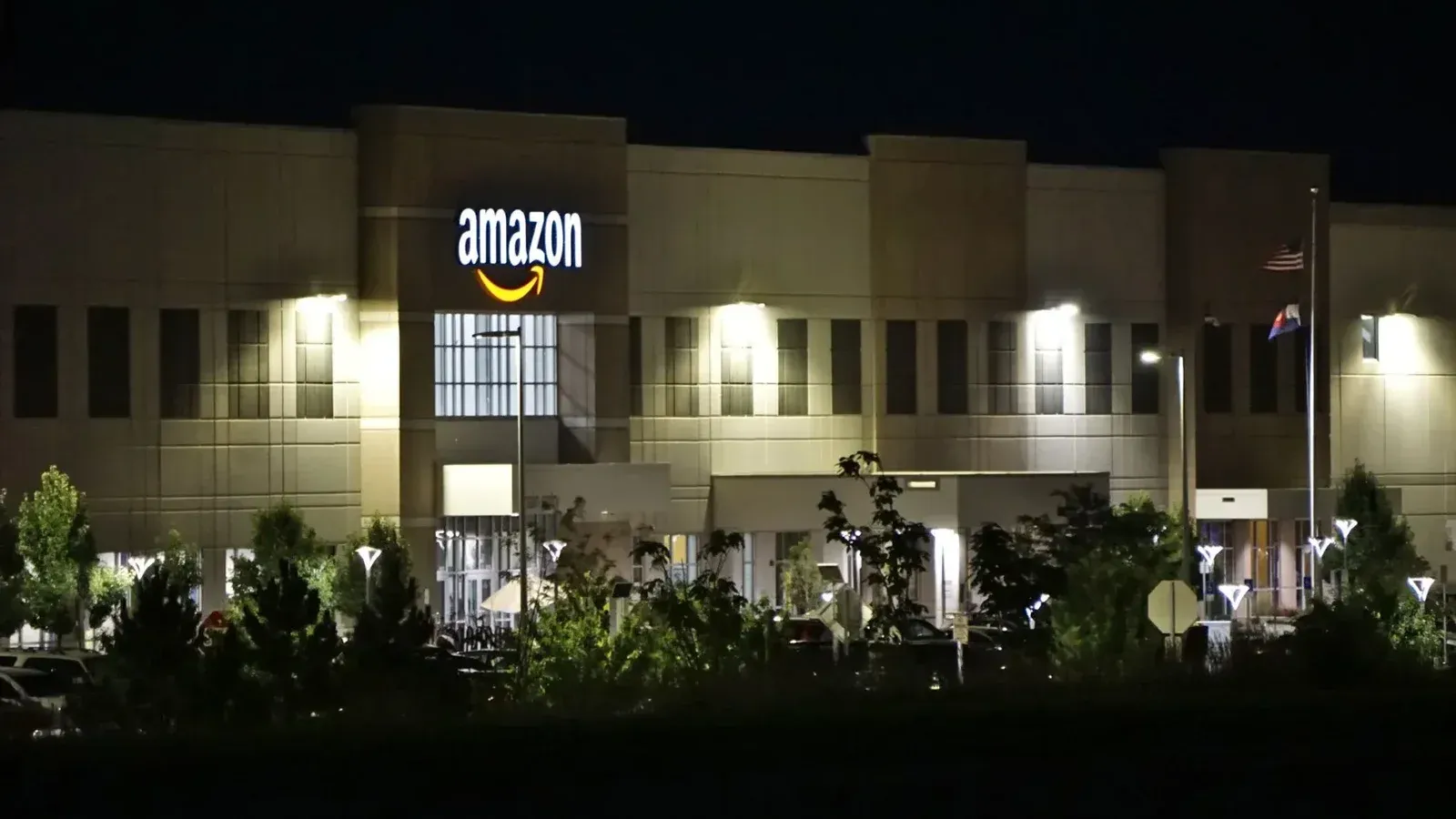 Edificio de almacén de Amazon iluminado por la noche con árboles y señalización.