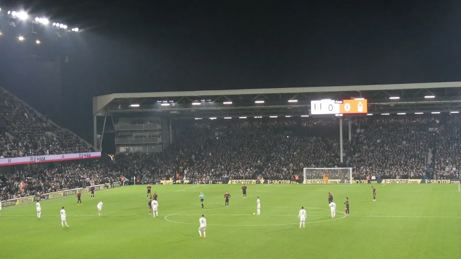 Vista nocturna del estadio del Fulham con las gradas llenas y los jugadores colocados en el campo bajo los focos, con el marcador al fondo.