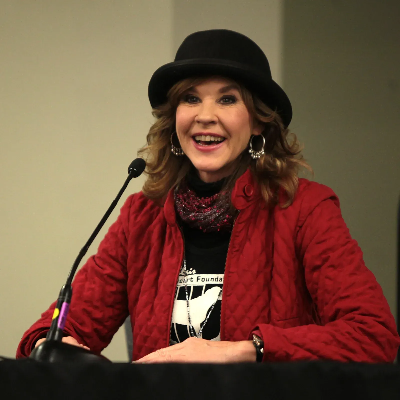 Linda Blair sonríe y habla ante un micrófono, con sombrero negro, chaqueta roja acolchada y una camiseta con la silueta de un perro.