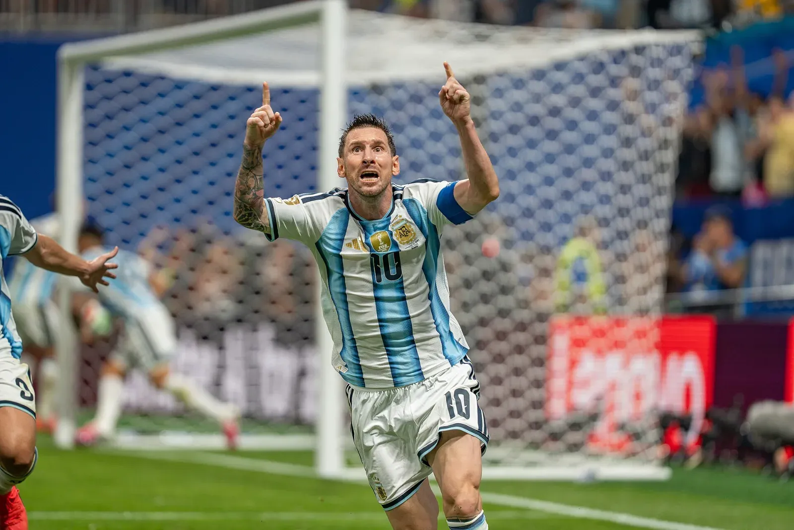 Lionel Messi, con la camiseta albiceleste número 10 y el brazalete de capitán, celebra con ambos índices hacia arriba frente a una portería.
