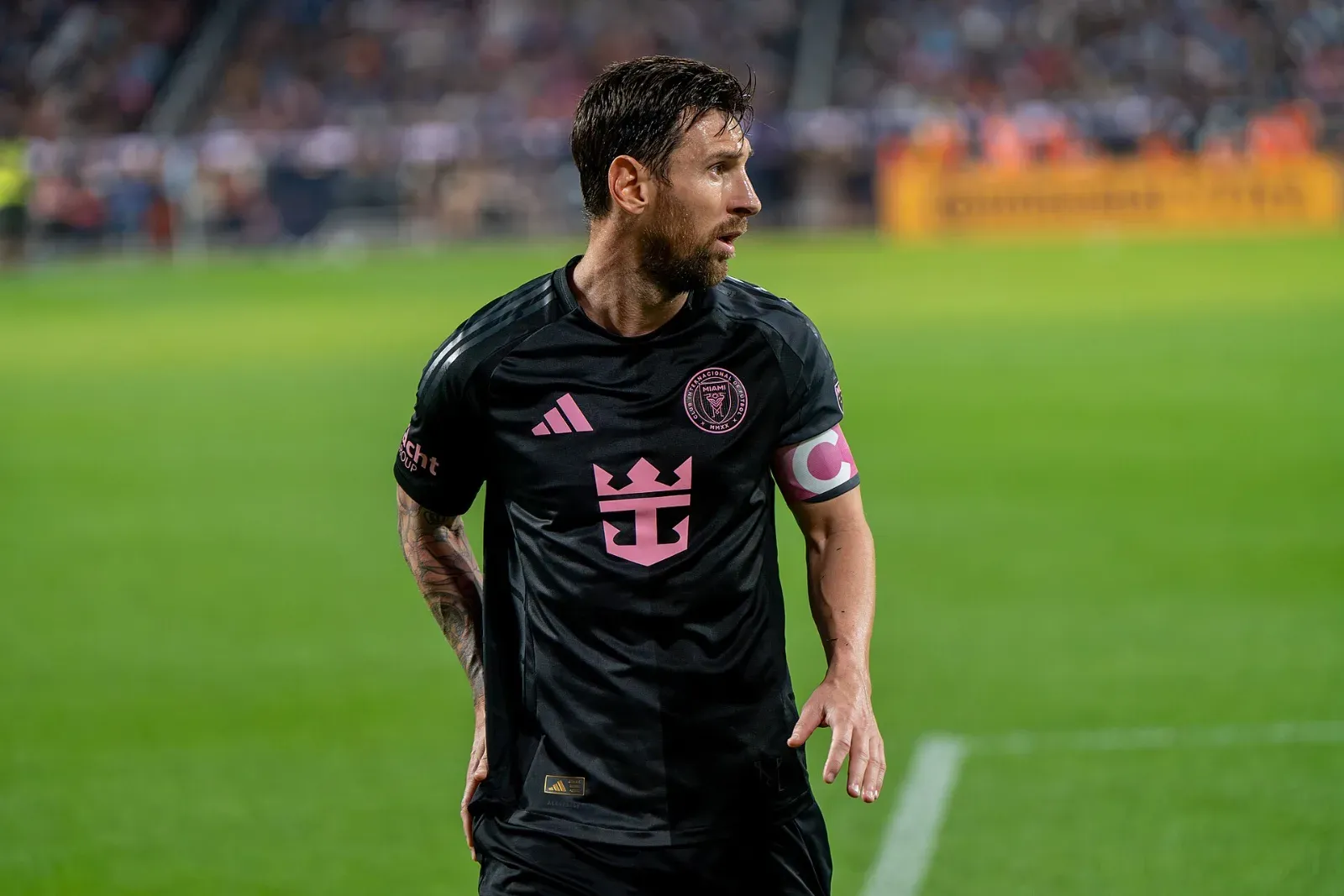 Lionel Messi, con la camiseta negra y rosa de Inter Miami y el brazalete de capitán, mira hacia un lado sobre el césped de un estadio.