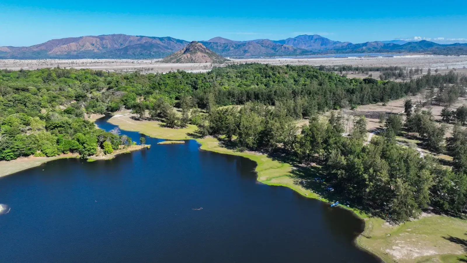 Breathtaking aerial view of a peaceful blue lake and lush green forest with mountain backdrop under clear blue skies.