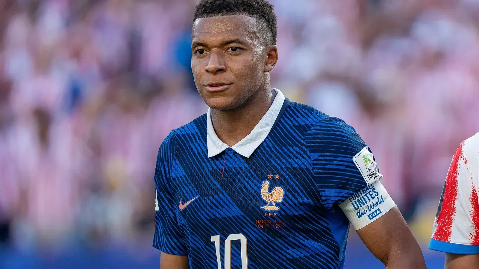 Kylian Mbappé, con la camiseta azul número 10 de Francia y el brazalete de capitán, sobre el campo durante el partido ante Paraguay; al fondo, aficionados con camisetas rojas y blancas.