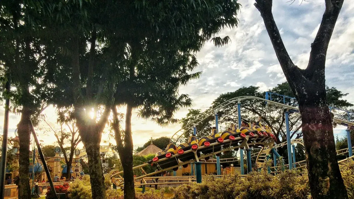 Escena vibrante de parque de atracciones con montaña rusa entre árboles exuberantes y hierba a la luz del día.